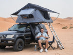 Load image into Gallery viewer, DesertX Nomad large grey rooftop tent set up on a black car in a desert landscape. Includes ladder and seating area.