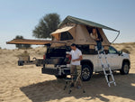 Load image into Gallery viewer, DesertX Nomad rooftop tent on a white truck, tan and green canvas, with a woman inside and a man on a ladder. Features an extendable awning.