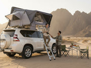 DesertX Basecamp grey hardshell rooftop tent set up on a white SUV, with a couple preparing for an outdoor adventure in a desert landscape.