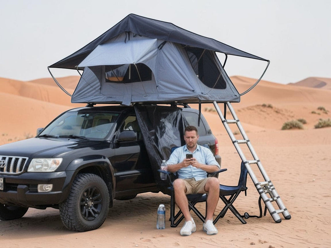 DesertX Nomad large grey rooftop tent set up on a black car in a desert landscape. Includes ladder and seating area.