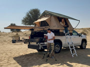 DesertX Nomad rooftop tent on a white pickup truck in a sandy desert landscape. Features a tan tent with a green rainfly and an attached awning. A couple is inside and near the tent.