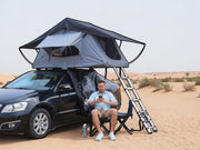 DesertX Nomad grey rooftop tent on black car in desert landscape with man sitting in chair.